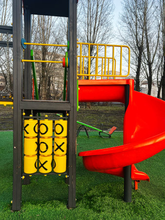A vibrant outdoor playground featuring a tic-tac-toe game and a curving red slide, set against a...