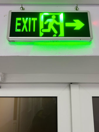 Green illuminated exit sign displaying a running figure with an arrow, indicating an emergency exit located above a doorway. Useful for safety demonstrations, emergency planning guides, and evacuation instructions.の写真素材