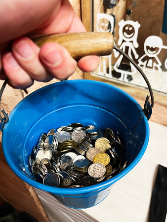 A close-up of a blue bucket containing mixed coins, symbolizing savings, wealth, or charity. The bucket is detailed with a wooden handle, offering a sense of tradition and tangibility in financial contexts.の写真素材