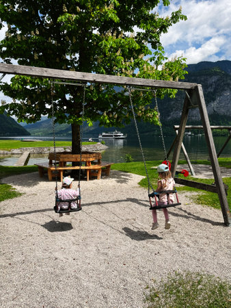 Children swing on wooden frames beside a beautiful lake under a clear sunny sky, Hallstatt, Austria. The surrounding scenery features lush greenery, a calm lake, and a peaceful environment perfect for relaxation.の写真素材