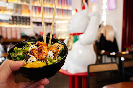 Hand holding a poke bowl in a lively restaurant featuring modern Asian style interior decorの写真素材