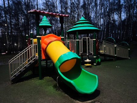 Playground with multicolored slides surrounded by trees under evening lighting.