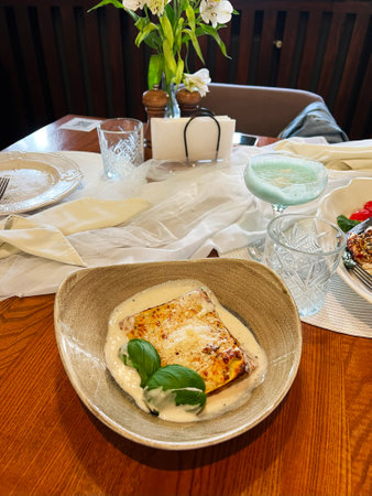 A beige bowl of lasagna in creamy sauce on a well-decorated restaurant table settingの写真素材