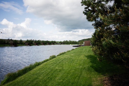 Picturesque riverbank landscape with green grass, trees, and wooden cabins on a cloudy day