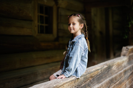 Child with braided hair wearing denim jacket seated near rustic wooden buildingの写真素材