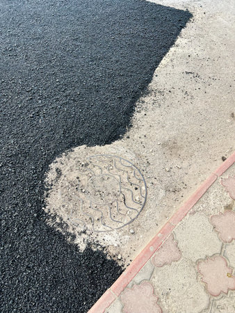 Fresh asphalt pavement contrasts with a decorative sidewalk and visible utility manhole cover.の写真素材