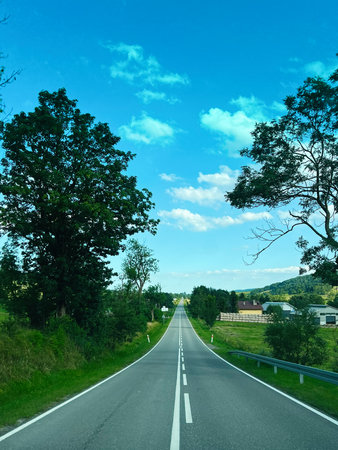 Sunny countryside route surrounded by green trees, open fields, and distant rural houses.の写真素材