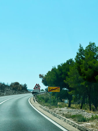 Curved Zaboric, Croatia road, greenery, and transportation signs under an expansive blue skyの写真素材