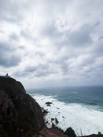 Coastal cliffs and lighthouse along the Cape Roca coast under overcast skies, with waves crashing below.の写真素材