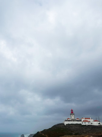 Serene coastal scene with a lighthouse, white structures, and red roofs under a dramatic, cloudy sky.  Cabo da Roca Lighthouseの写真素材