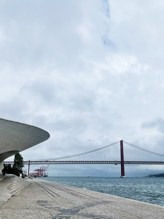 Lisbon waterfront scene featuring the MAAT curved facade and the red suspension bridge over the Tagus.の写真素材