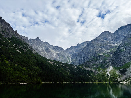 Serene alpine lake reflects towering cliffs and forested slopes in Zakopane, Poland under a dramatic sky.の写真素材