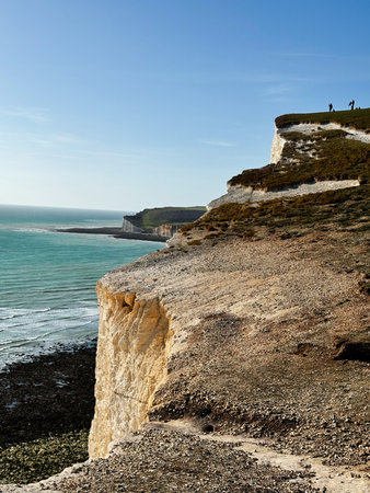 Rugged chalk cliffs meet blue sea under clear skies at Seven Sisters, with hikers on the edge.