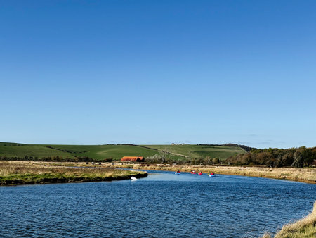 Kayakers paddle along a calm river with green fields and a clear blue sky.