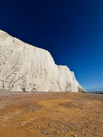 White chalk cliffs rise above a brown pebble beach under a clear blue sky on the East Sussex coast.