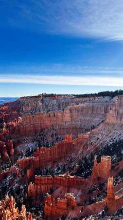 A dramatic Utah canyon scene featuring red hoodoos, blue sky, snow, and pine forests. Bryce Canyon National Parkの写真素材