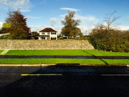 Longford, Ireland - October 21, 2025: A peaceful Irish street view with a house beyond a stone wall, lush grass, and clear blue sky.のeditorial素材