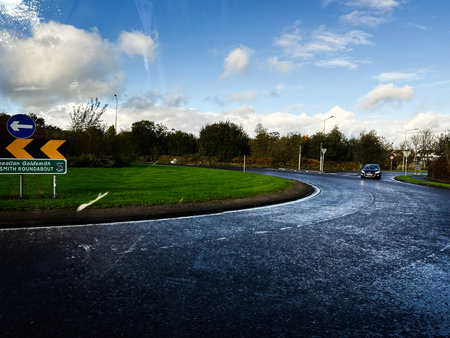 Edgeworthstown, Ireland - October 21, 2025: Curved Goldsmith roundabout in Ireland with a car on a wet road beneath a bright sky and lush greenery.のeditorial素材