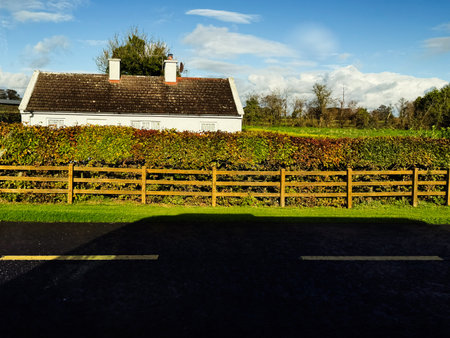 Longford, Ireland - October 21, 2025: Bright rural scene in Ireland with a white cottage, lush hedge, and wooden fence under blue skies.のeditorial素材