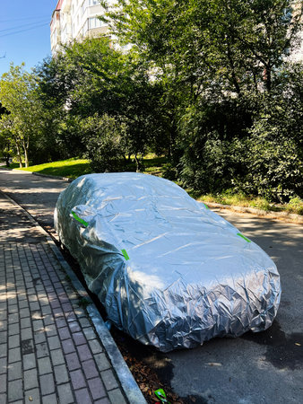 A silver car is fully covered with a reflective tarp on a calm tree lined street in a residential area.の写真素材