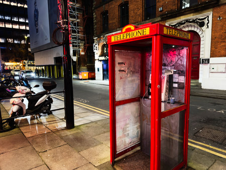 Manchester, UK - December 10, 2025: Bright red telephone booth on a Manchester street at night beside parked scooters and brick buildings.のeditorial素材