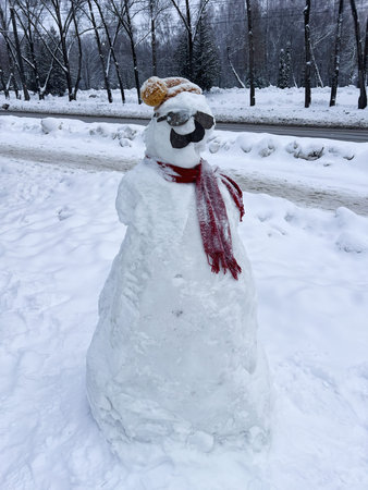 A playful snowman with a red scarf and sunglasses stands proudly in a snowy park.の写真素材
