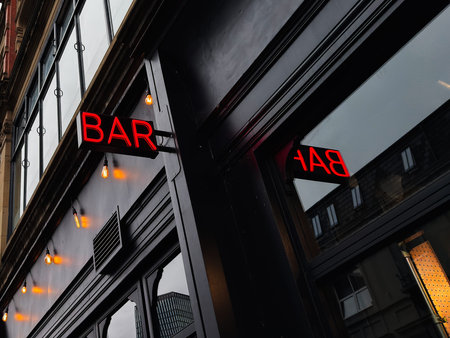 Bar neon sign on a dark storefront in Manchester, UK, viewed from below against a cloudy sky.