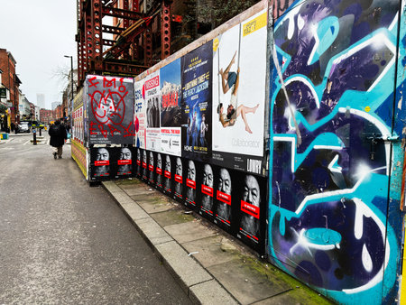 Manchester, UK - January 30, 2026: People walk past a poster covered wall with graffiti in...