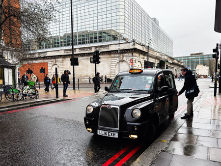 London, UK - February 01, 2026: A classic London black taxi waits on a rainy, urban street with pedestrians nearby.のeditorial素材