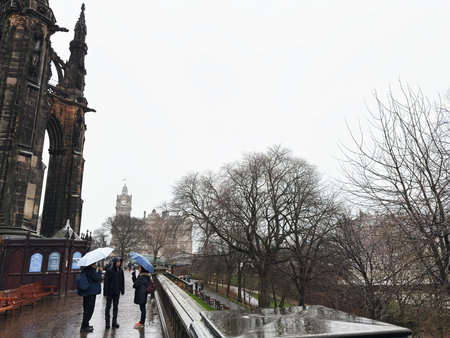 Edinburgh, Scotland, UK - February 15, 2026: Three people under umbrellas stand by the gothic Scott...