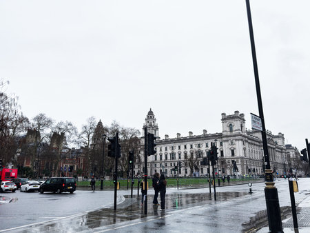 London, UK - February 01, 2026: Pedestrians wait at traffic lights on a wet street beside a large...