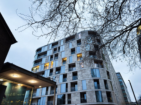 Dusk view of a modern curved apartment building in London, UK with illuminated windows and bare tree...