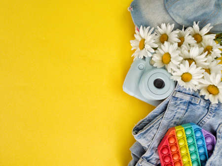 On a bright yellow background are jeans, a baseball cap, a camera, a popit toy and a bouquet of daisies. High quality photoの写真素材