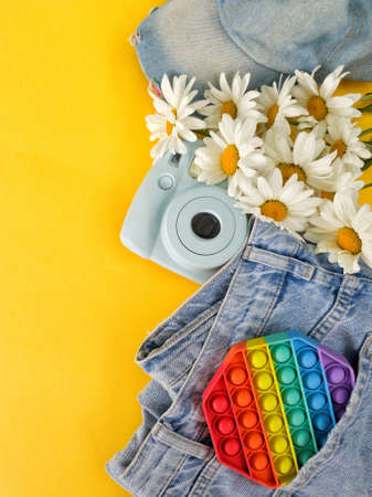 On a bright yellow background are jeans, a baseball cap, a camera, a popit toy and a bouquet of daisiesの写真素材