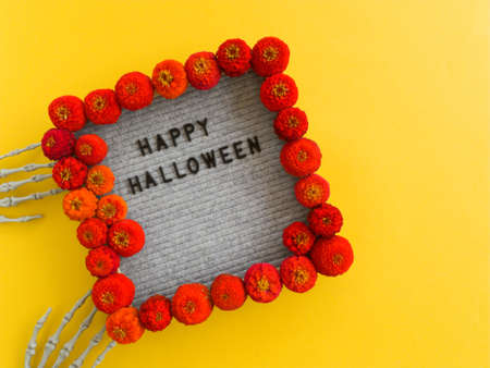 The word Halloween is framed in the hands of a skeleton and framed with red flowers on a yellow...