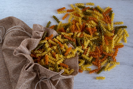 Uncooked multicolored Fusilli pasta spilled out of fabric bag over white wood background. top view.の写真素材
