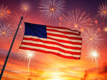 A vivid image of the American flag waving against a backdrop of a colorful fireworks display at dusk. The flag is prominently featured with its stars and stripes clearly visible, while the fireworks burst in various colors and patterns in the sky.の素材