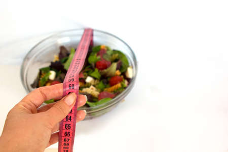 female hand holding a pink centimeter at 60 centimeters against a transparent glass bowl with salad, tomatoes, mozzarella, salami, pasta on a white backgroundの写真素材