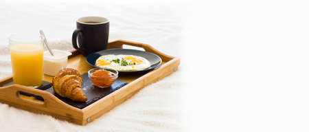 Wooden coffee tray with fried eggs, croissant with jam, cup of tea, juice and yogurt on white bedding. Breakfast in the bed good morning concept. Romantic Weekend. Vacations.Banner. Space for textの写真素材