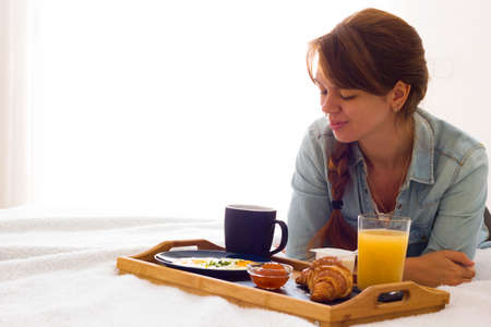 Happy girl having breakfast lying on the bed and looking at the coffee tray. Space for text. Good morning surprise. Breakfast in bed conceptの写真素材
