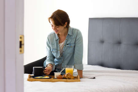 opened white door to bedroom interior with relaxed happy woman having breakfast in bed. Big...