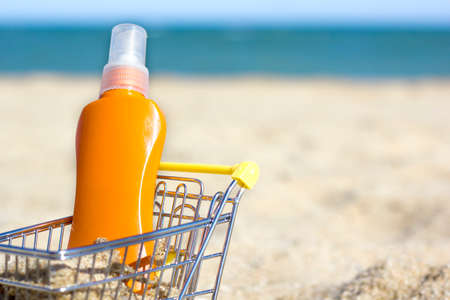 Close up a orange bottle of sunscreen in a Grocery Shopping Cart on the beach sea and blue sky background. Holiday shopping. spf scik care. Protect your skin. Space for text. Summertime. Mock up.の写真素材