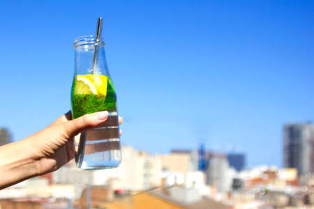 woman hand holding a bottle with refreshing drink with water, lemon and mint with a reusable metal drinking straw on city background. Happy zero waste summerの写真素材