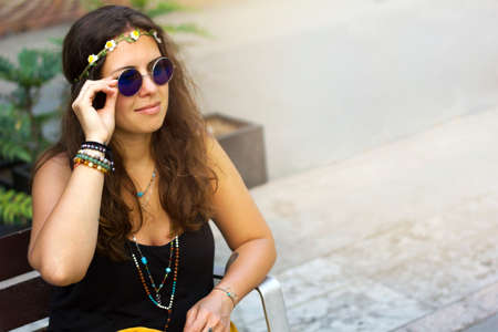 Beautiful young curly hair hippie woman with rounded vontage blue sunglasses sitting on a bench in the squareの写真素材