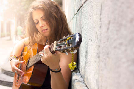 Cheeful curly brunette girl with vintage blue sunglasses and guitar sitting in the square and looking up at the sky and sun. Hippie free lifestyle. Street musician. Happy and relaxの写真素材