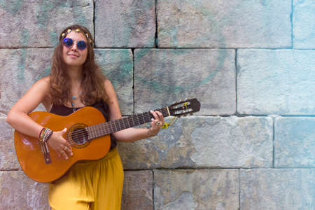Curly hair hippie young woman with rounded blue vintage sunglasses smiling and playing acoustic guitar on the street. Free and happy lafestyle. Enjoy the music.の写真素材