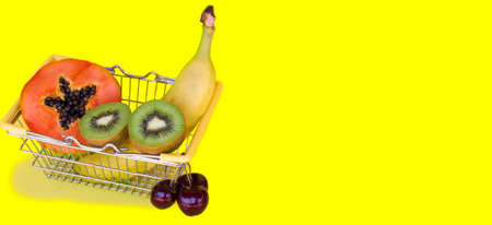 Shopping basket full of tropical exotic fruits on yellow background with space for text. Sweet Papaya, banana, kiwi and cherry. Shopping online. Fresh fruit fast delivery. Buy healthy food.の写真素材
