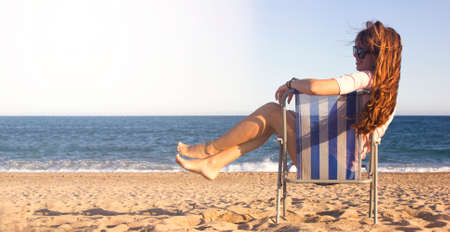 Summer beach vacation concept, cheerful young woman with sunglasses with long ginger hair smiling and relaxing on chair beach with sea on background. Space for text. Girl on beach in Spainの写真素材