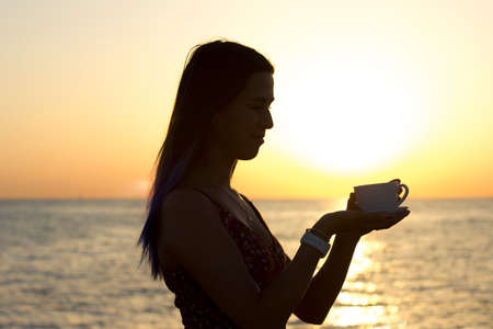 silhouette of a young girl with long hair holding a cup of tea or coffee at sunrise on the beach...
