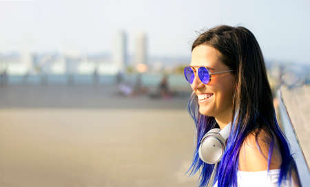 Close - up portrait of a young beautiful happy girl with blue hair in sunglasses and with big headphones. Informal style. Street portrait of cheerful woman. summer pleasuresの写真素材